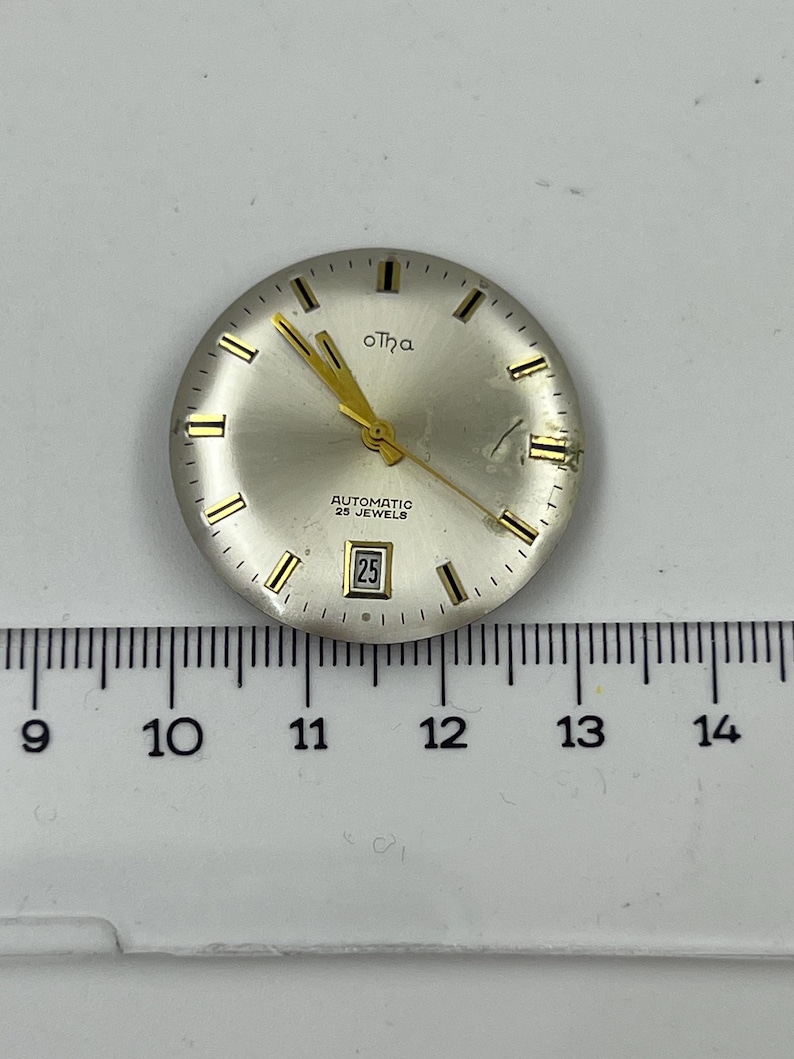 May include: A silver watch face with gold hands and markers. The watch face has the text "oTha", "AUTOMATIC 25 JEWELS", and a date display showing "25". A ruler is below the watch face.
