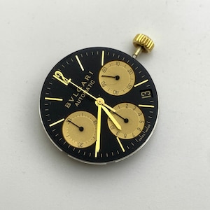 May include: A black watch face with gold accents, featuring the brand name "BVLGARI AUTOMATIC". It has three sub-dials, gold hands, and a date display. The watch face includes the text "SWISS MADE".