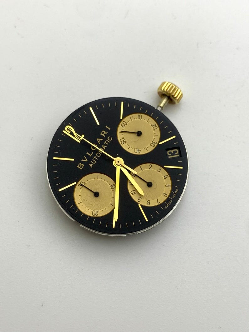 May include: A black watch face with gold accents, featuring the brand name "BVLGARI AUTOMATIC". It has three sub-dials, gold hands, and a date display. The watch face includes the text "SWISS MADE".