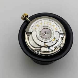 May include: A close-up of a watch movement with the brand name "BVLGARI" in gold lettering. The watch mechanism is silver with intricate gears and a gold crown. The movement is set in a black holder.