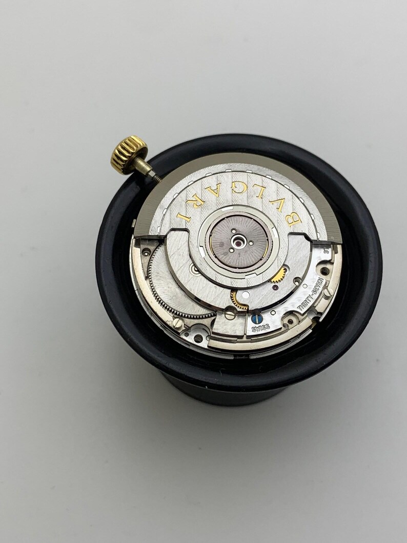May include: A close-up of a watch movement with the brand name "BVLGARI" in gold lettering. The watch mechanism is silver with intricate gears and a gold crown. The movement is set in a black holder.