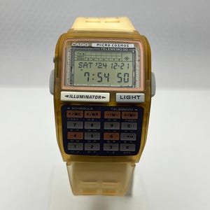 Può includere: Un orologio digitale vintage Casio Micro Cosmos Telememo 50. L'orologio ha una tastiera di calcolatrice e un display digitale che mostra la data e l'ora. L'orologio è racchiuso in plastica gialla traslucida con un cinturino abbinato.