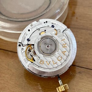 May include: Close-up of a watch movement with intricate gears and components. The silver-toned mechanism features gold accents and the brand name "BVLGARI" in capital letters. A gold-colored crown is attached to the side. The watch movement is on a wooden surface.