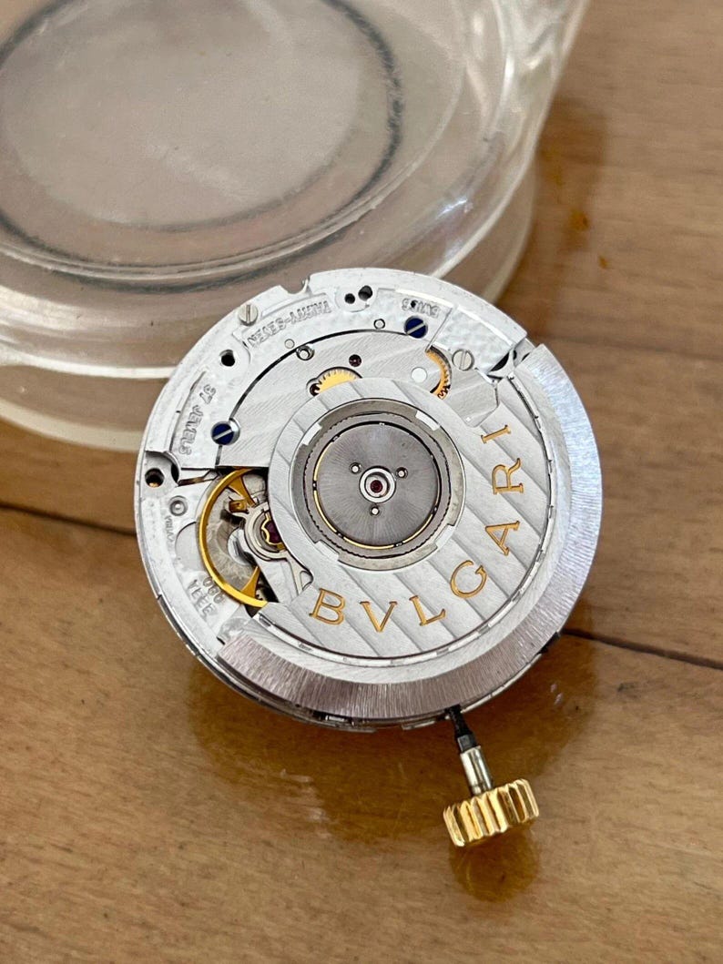 May include: Close-up of a watch movement with intricate gears and components. The silver-toned mechanism features gold accents and the brand name "BVLGARI" in capital letters. A gold-colored crown is attached to the side. The watch movement is on a wooden surface.