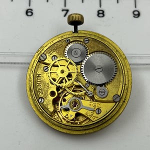May include: A close-up of a vintage Zenith watch movement. The gold-toned mechanism features intricate gears, screws, and a winding stem. The word "ZENITH" is visible on the face, along with the serial number 678823.