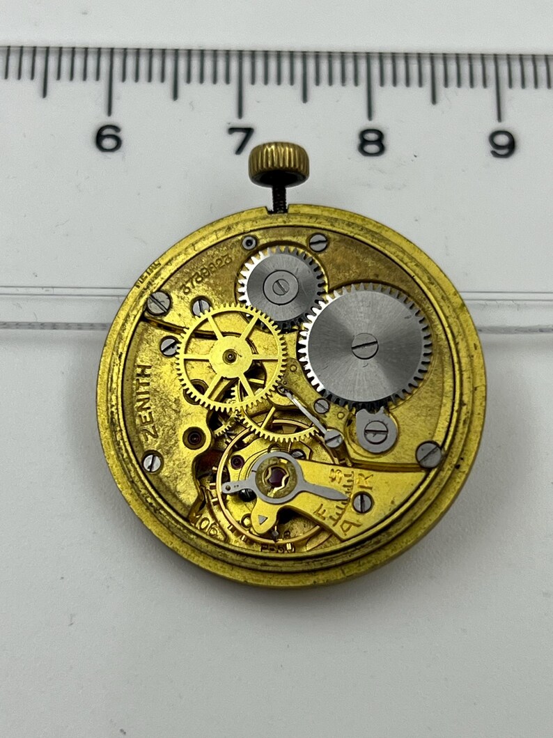 May include: A close-up of a vintage Zenith watch movement. The gold-toned mechanism features intricate gears, screws, and a winding stem. The word "ZENITH" is visible on the face, along with the serial number 678823.