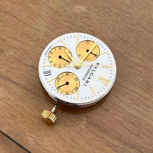 May include: A white watch face with gold accents and three sub-dials. The watch face has the text "BVLGARI AUTOMATIC" and the number "11". The watch face has a gold crown.