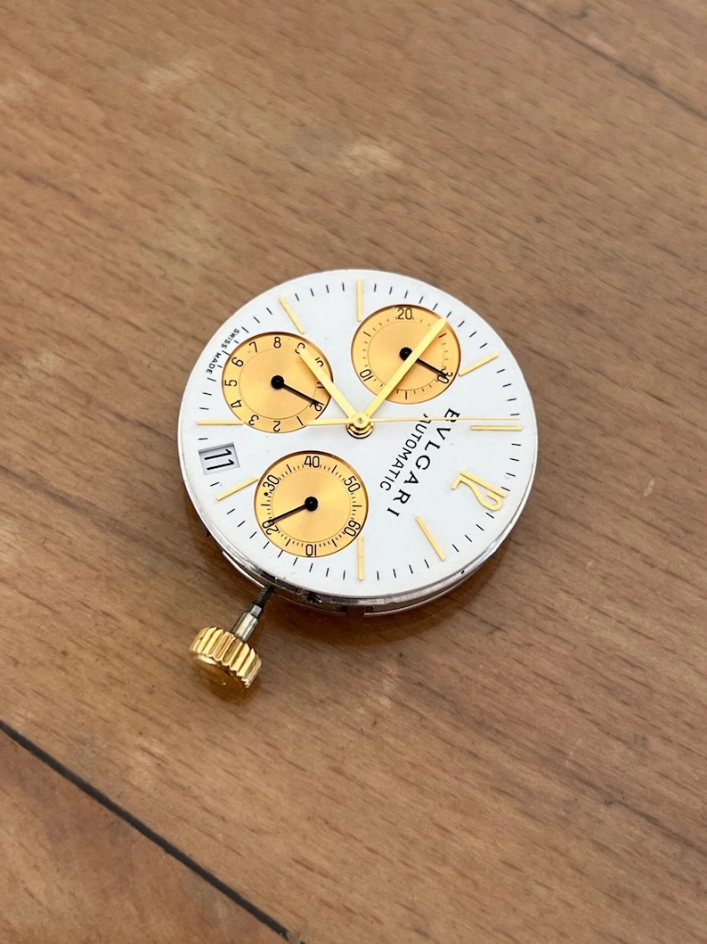 May include: A white watch face with gold accents and three sub-dials. The watch face has the text "BVLGARI AUTOMATIC" and the number "11". The watch face has a gold crown.