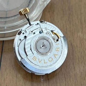 May include: Close-up of a watch movement with the brand name "BVLGARI" in gold lettering. The movement has intricate gears, a gold-colored crown, and various silver-colored components. The model number "58505" is visible.