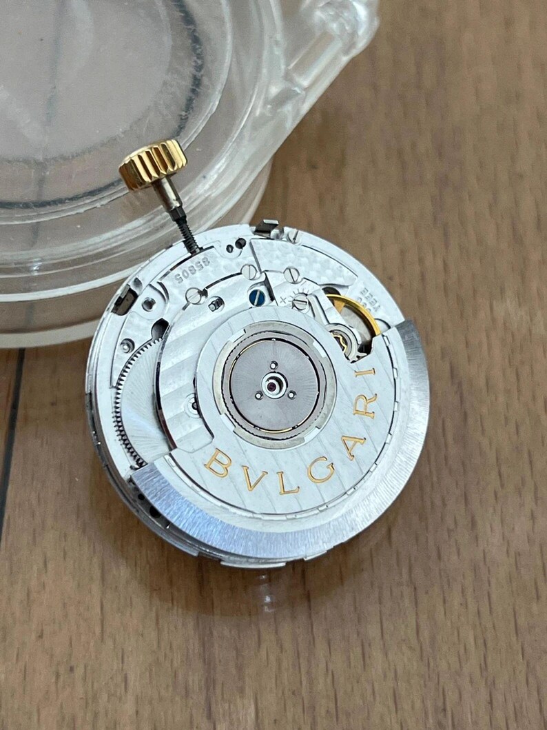 May include: Close-up of a watch movement with the brand name "BVLGARI" in gold lettering. The movement has intricate gears, a gold-colored crown, and various silver-colored components. The model number "58505" is visible.