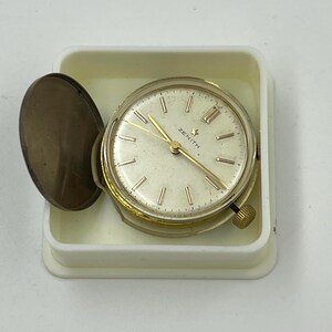 May include: A vintage Zenith watch with a gold-tone case and hands, set in a white plastic container. The watch face is cream-colored with Roman numerals and the Zenith logo. The watch has a small winding crown and a hinged cover.