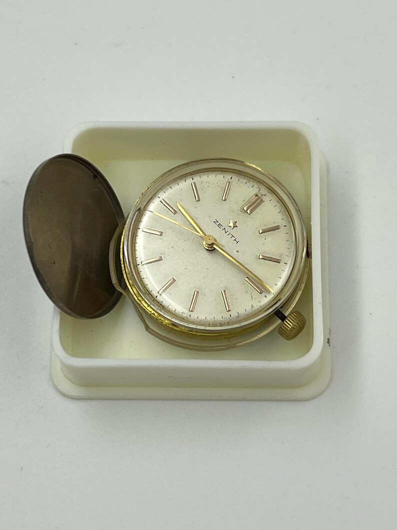 May include: A vintage Zenith watch with a gold-tone case and hands, set in a white plastic container. The watch face is cream-colored with Roman numerals and the Zenith logo. The watch has a small winding crown and a hinged cover.