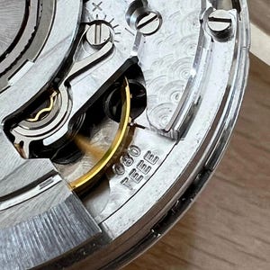 May include: Close-up of a watch movement, showcasing intricate mechanical components. Silver-toned gears, screws, and springs are visible, with a gold-colored balance wheel and a small inscription. The image highlights the precision engineering of a timepiece.