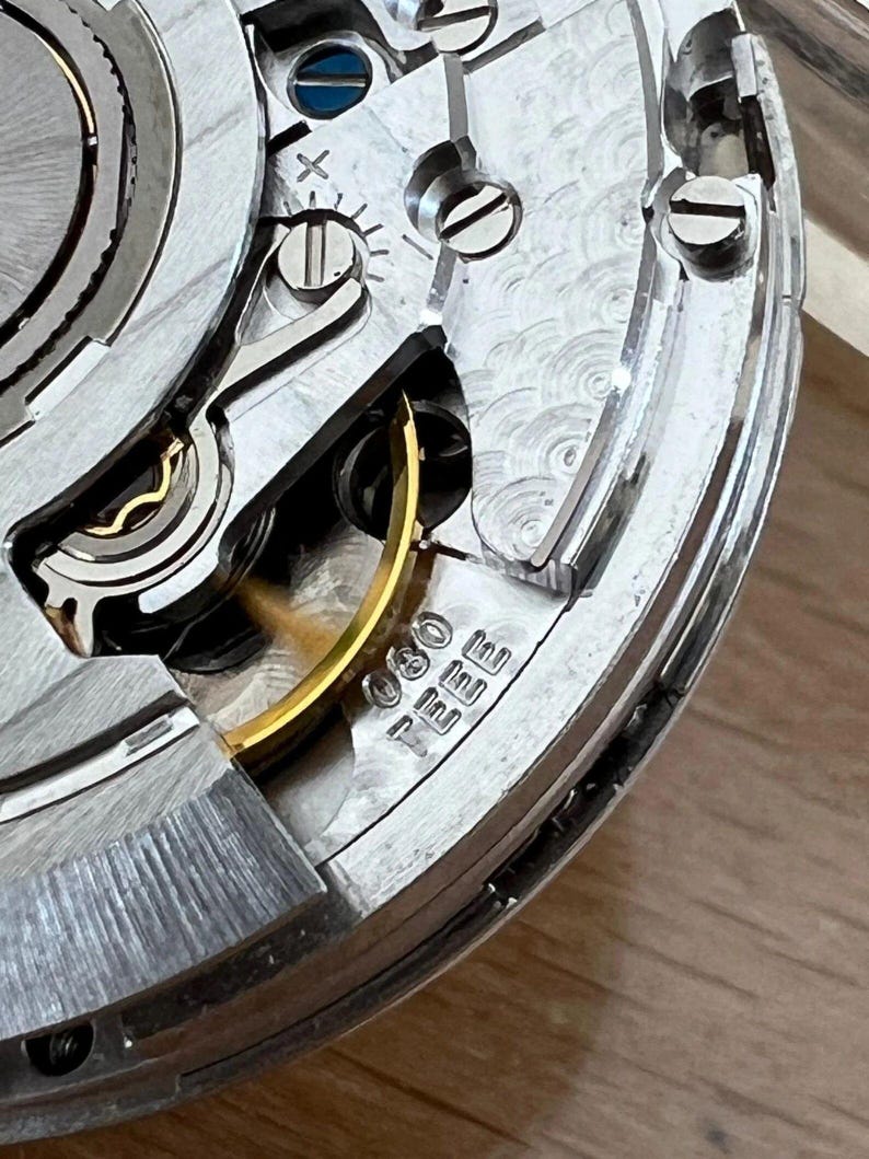 May include: Close-up of a watch movement, showcasing intricate mechanical components. Silver-toned gears, screws, and springs are visible, with a gold-colored balance wheel and a small inscription. The image highlights the precision engineering of a timepiece.