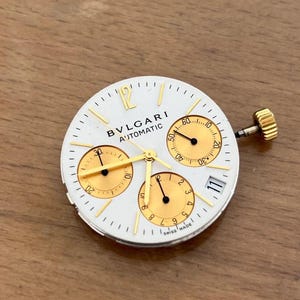 May include: A white watch face with gold accents and the text "BVLGARI AUTOMATIC". The watch face has three sub-dials, gold hands, and a gold crown. The date is displayed at the 3 o'clock position. Swiss Made.