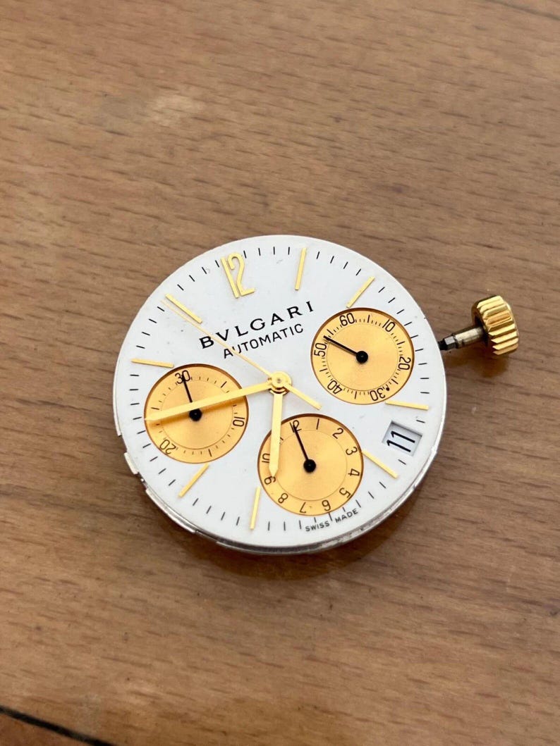 May include: A white watch face with gold accents and the text "BVLGARI AUTOMATIC". The watch face has three sub-dials, gold hands, and a gold crown. The date is displayed at the 3 o'clock position. Swiss Made.