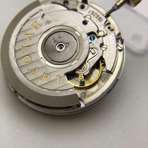 May include: Close-up of a watch movement with the brand name "BVLGARI" in gold lettering. The intricate mechanism features various gears, screws, and a gold-colored crown. The watch movement is silver and gold in color.