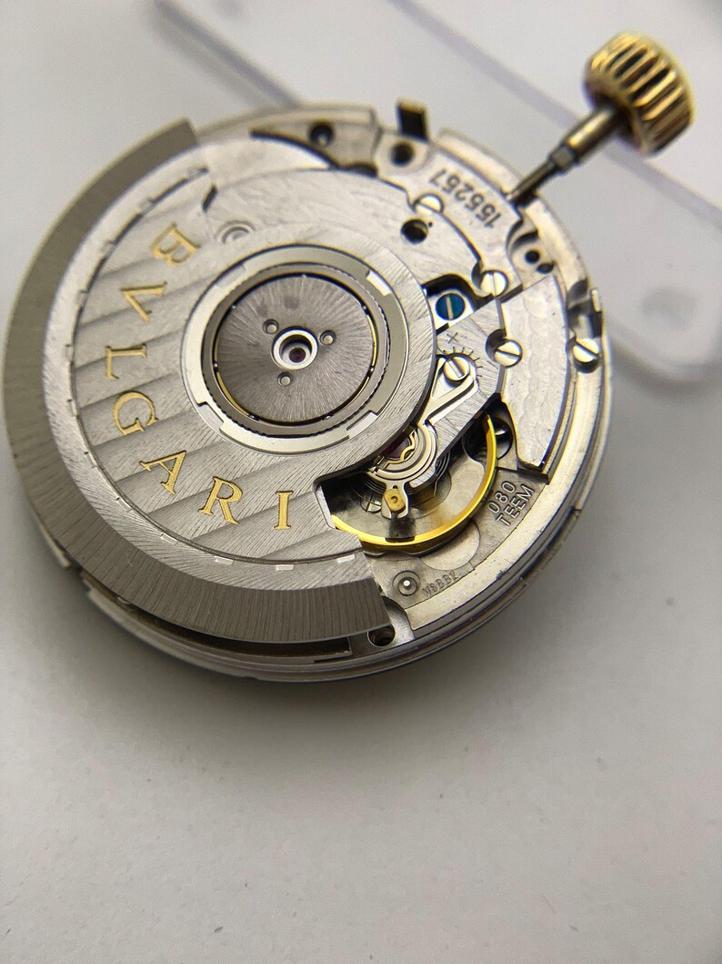 May include: Close-up of a watch movement with the brand name "BVLGARI" in gold lettering. The intricate mechanism features various gears, screws, and a gold-colored crown. The watch movement is silver and gold in color.