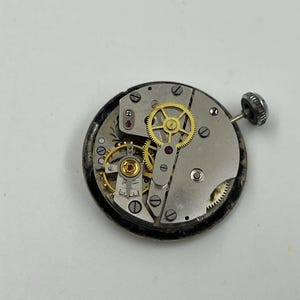 以下が含まれることがあります： 歯車、ネジ、宝石が見える時計のムーブメントのクローズアップ。金属部品は銀色と金色で、外側のリングは黒色です。時計のステムとリューズが右側にあります。