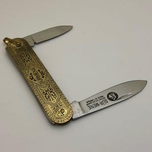 Può includere: Un piccolo coltello tascabile aperto con due lame. Il manico è color oro con intricati disegni neri incisi e la parola "Sürmene". Una lama è aperta e l'altra è parzialmente aperta. La lama ha il testo "SÜR-MENE" e un logo.