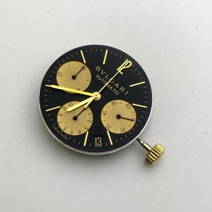May include: A black watch face with gold accents, featuring three sub-dials and a date window displaying the number 13. The watch face has the brand name "BVLGARI AUTOMATIC" printed on it. The watch has a gold crown.