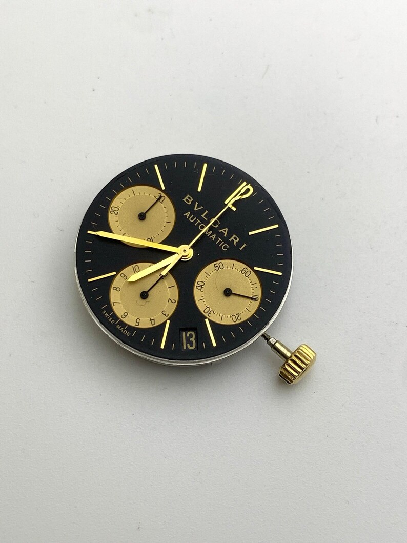 May include: A black watch face with gold accents, featuring three sub-dials and a date window displaying the number 13. The watch face has the brand name "BVLGARI AUTOMATIC" printed on it. The watch has a gold crown.