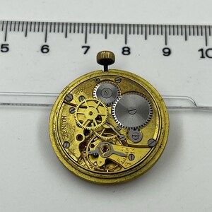 May include: Close-up of a vintage Zenith watch movement. The gold-toned mechanism features intricate gears, a balance wheel, and a winding stem. The watch movement is set against a white background with a ruler for scale.