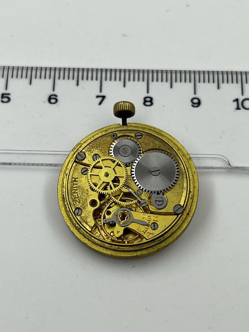 May include: Close-up of a vintage Zenith watch movement. The gold-toned mechanism features intricate gears, a balance wheel, and a winding stem. The watch movement is set against a white background with a ruler for scale.