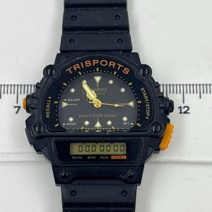 May include: Black Casio Trisports digital watch with a black band. The watch face displays the text "TRISPORTS", "CASIO", and "WATER 50M RESIST". The digital display shows "000 00 00". Orange accents are visible on the watch.