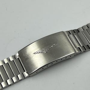 May include: A stainless steel watch strap with a brushed finish. The clasp features a logo with a winged hourglass design. The strap is made up of multiple rectangular links.