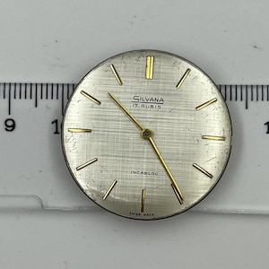 Könnte beinhalten: Ein Vintage-Zifferblatt mit silberfarbenem Zifferblatt und goldfarbenen Zeigern und Markierungen. Das Zifferblatt trägt den Markennamen "SILVANA" und den Text "17 RUBIS" und "INCABLOC". Die Worte "SWISS MADE" sind ebenfalls sichtbar.