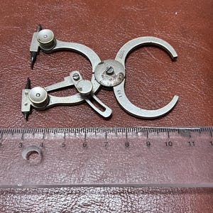 May include: A vintage silver-toned caliper tool with curved arms and pointed tips. The tool has a circular dial and is marked with the number 199. A clear ruler with centimetre markings is in the foreground.