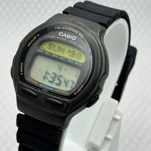 Puede incluir: Un reloj digital Casio Telememo 50 con correa negra y caja gris. La esfera del reloj muestra la hora, el día y la fecha. El reloj tiene botones para ajustar la hora y otras funciones. El reloj se muestra sobre un soporte blanco.