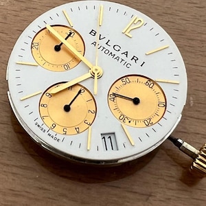 May include: Close-up of a white watch face with gold accents and three sub-dials. The watch face has the text "BVLGARI AUTOMATIC" and "SWISS MADE". The date is displayed as "11". Gold hands point to the time.