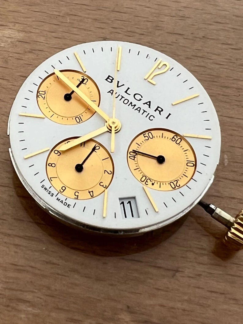 May include: Close-up of a white watch face with gold accents and three sub-dials. The watch face has the text "BVLGARI AUTOMATIC" and "SWISS MADE". The date is displayed as "11". Gold hands point to the time.