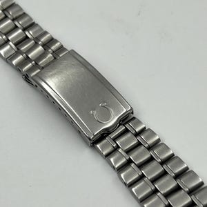 Pode incluir: Close-up de uma pulseira de relógio em aço inoxidável. A pulseira apresenta uma série de elos retangulares e um fecho com o logótipo Omega. O metal tem um acabamento escovado e a pulseira está angulada sobre um fundo branco.