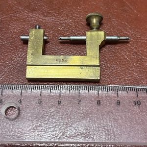 May include: A vintage brass tool with a U-shaped frame, adjustable arms, and a knurled knob. The tool has a small inscription, "1826", and is positioned next to a ruler. The tool appears to be a measuring instrument.