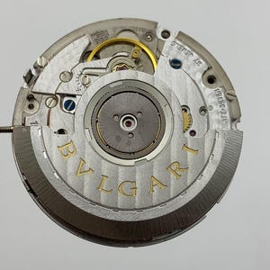 May include: Close-up of a watch movement with the brand name "BVLGARI" in gold lettering. The intricate mechanism features various gears, screws, and jewels, showcasing the precision of the watchmaking. The movement is silver and gold in color.