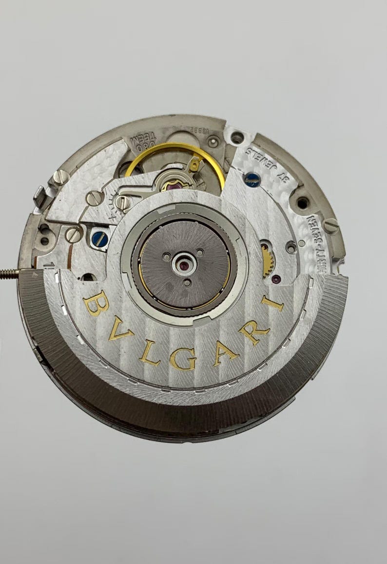 May include: Close-up of a watch movement with the brand name "BVLGARI" in gold lettering. The intricate mechanism features various gears, screws, and jewels, showcasing the precision of the watchmaking. The movement is silver and gold in color.