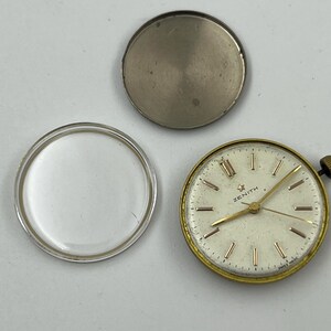 May include: An open vintage Zenith watch with a gold-tone bezel and a cream-colored face. The watch face has gold-tone hands and markers. The watch is disassembled, with the back and crystal separated. The watch face reads "ZENITH".
