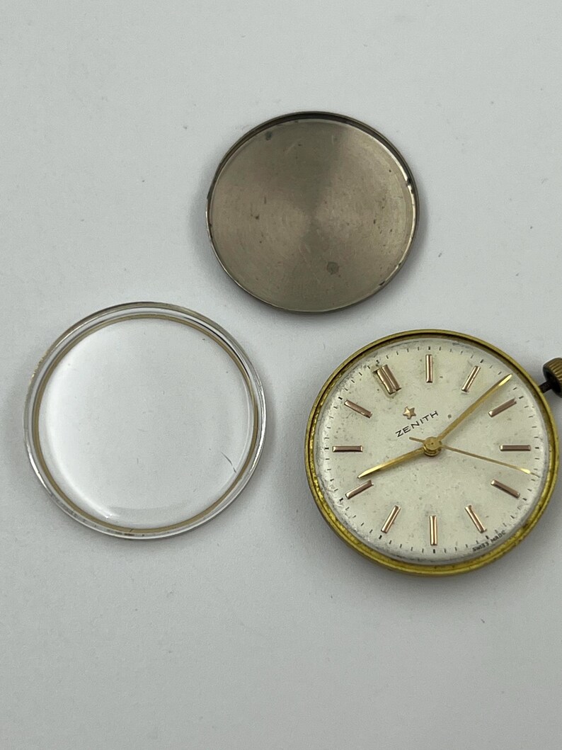 May include: An open vintage Zenith watch with a gold-tone bezel and a cream-colored face. The watch face has gold-tone hands and markers. The watch is disassembled, with the back and crystal separated. The watch face reads "ZENITH".