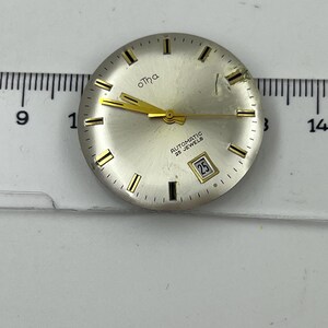 May include: A silver watch face with gold hands and markers. The watch face features the brand name "otha" in gold script and the text "AUTOMATIC 25 JEWELS". The date is displayed as "25" in a small window.