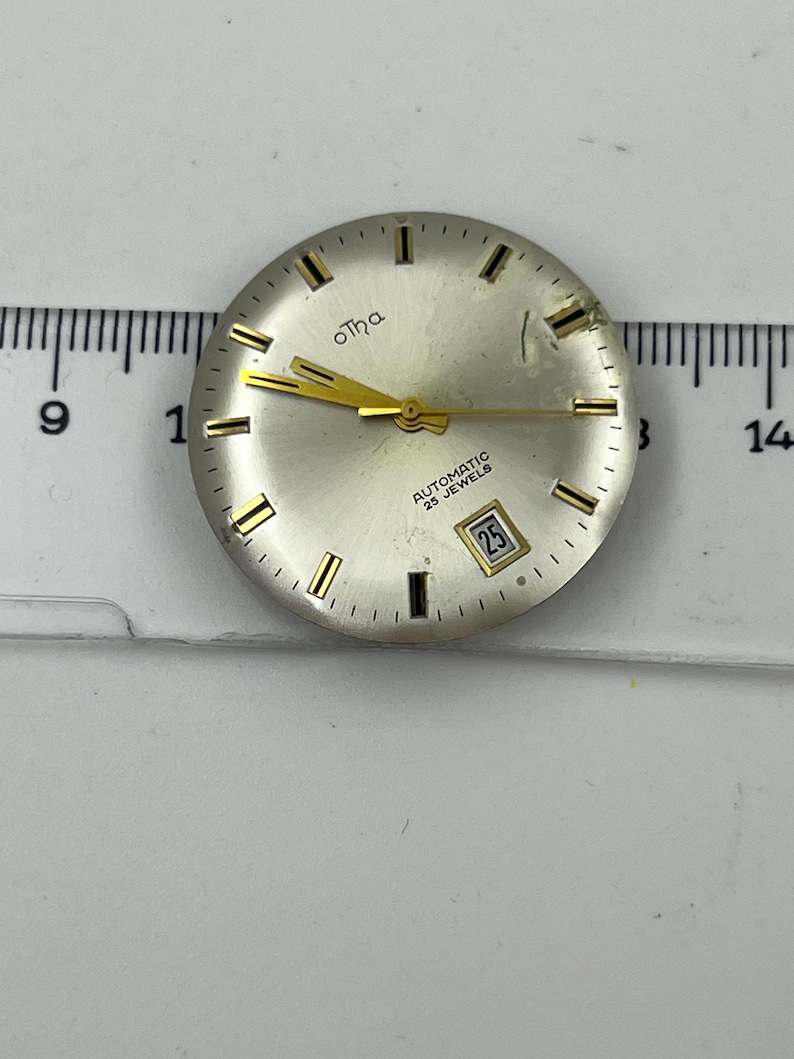 May include: A silver watch face with gold hands and markers. The watch face features the brand name "otha" in gold script and the text "AUTOMATIC 25 JEWELS". The date is displayed as "25" in a small window.