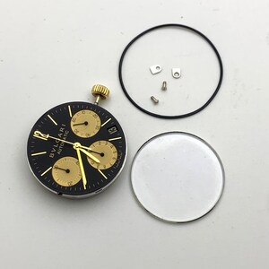 May include: A disassembled watch face with a black dial and gold accents. The watch face has three sub-dials and the text "BVLGARI AUTOMATIC". Other components include a crystal, a black gasket, and small screws.