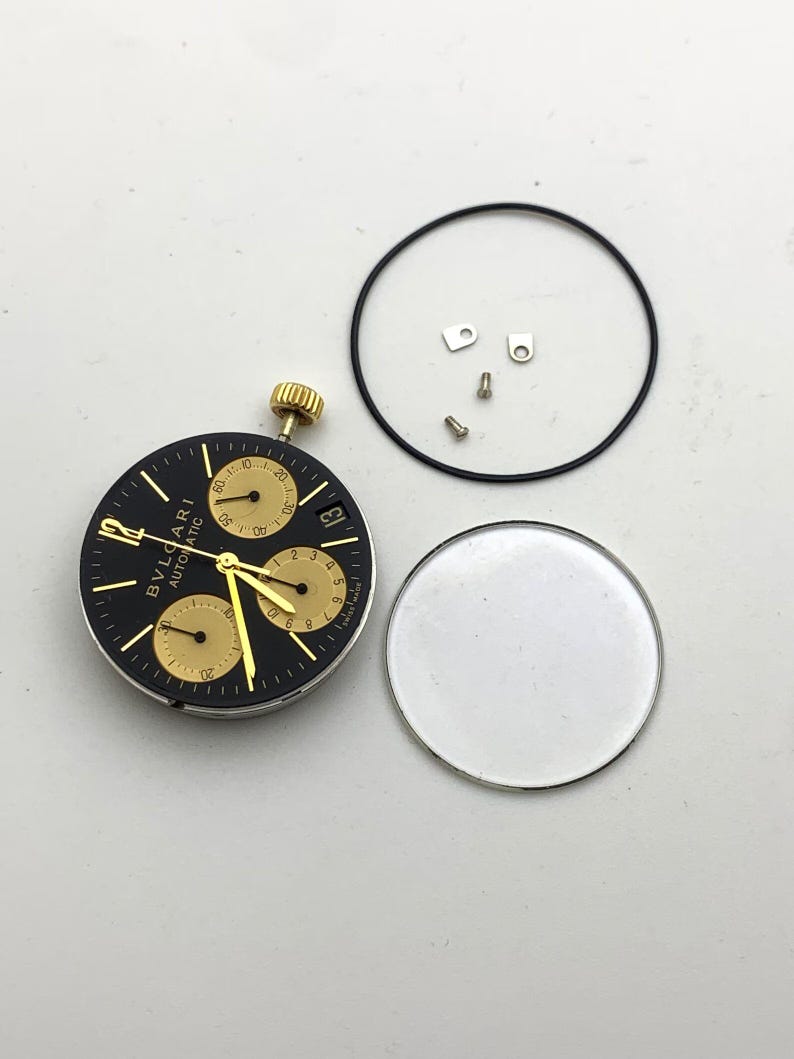 May include: A disassembled watch face with a black dial and gold accents. The watch face has three sub-dials and the text "BVLGARI AUTOMATIC". Other components include a crystal, a black gasket, and small screws.