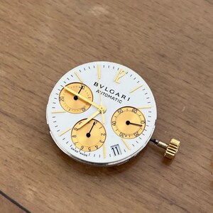 May include: White watch face with gold accents, featuring three sub-dials and the text "BVLGARI AUTOMATIC". The watch face has a date display at the 3 o'clock position and a gold crown.
