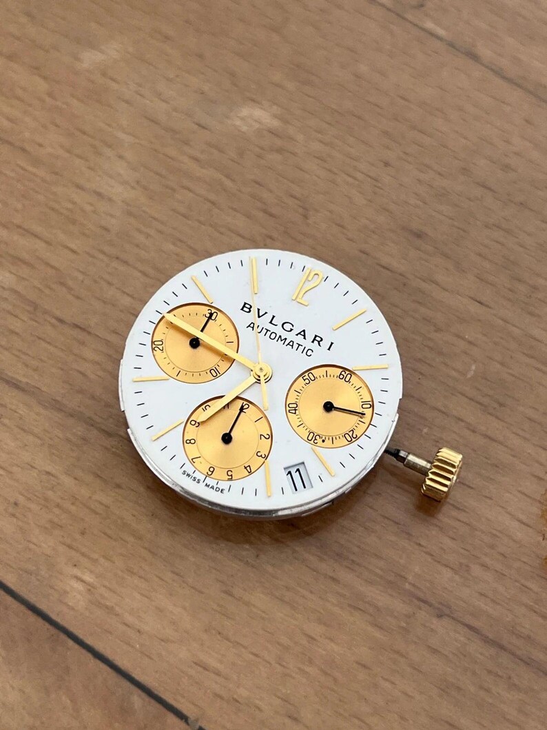 May include: White watch face with gold accents, featuring three sub-dials and the text "BVLGARI AUTOMATIC". The watch face has a date display at the 3 o'clock position and a gold crown.