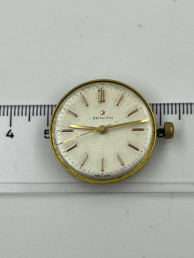 May include: Vintage Zenith watch face with a gold-tone bezel and hands. The off-white dial features gold markers and the Zenith logo. The watch face is approximately 1.25 inches in diameter and has a crown on the side. The words "SWISS MADE" are printed at the bottom.