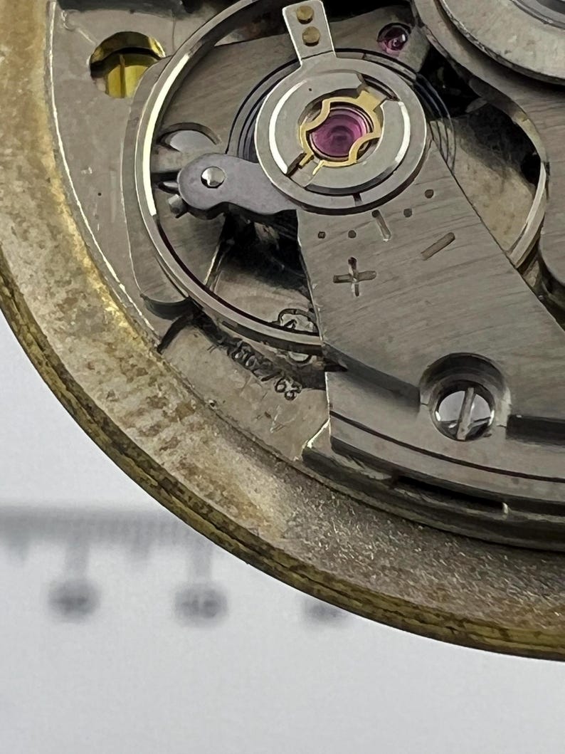 May include: Detailed view of a watch movement, revealing its complex internal workings. The image highlights metallic parts, such as gears, screws, and a balance wheel with a jewel. The serial number, 562/63, is stamped on the mechanism.