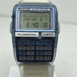 May include: A vintage Casio Micro Cosmos Telememo 50 digital watch. The watch has a blue and off-white case and band. The digital display shows the date, time, and day of the week. Features include an illuminator and light button.